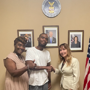 In front of the Department of Labor seal in a federal building, a man is shaking hands with a woman in a suit. Another woman stands beside him, giving a thumbs up to the camera. All are smiling.