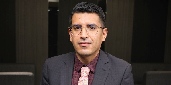 A man in a suit and tie sits at a table in a conference room.