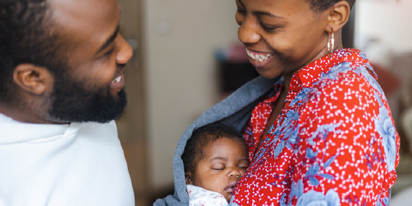 A happy family. A smiling woman cradles a small infant in a sling, standing with a smiling man.