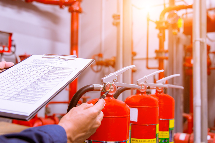 A worker holds a clipboard while inspecting fire extinguishers in an industrial environment