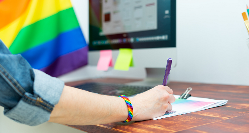 A worker wearing a rainbow bracelet writes on a piece of paper at a desk. A Pride flag is displayed next to their computer.
