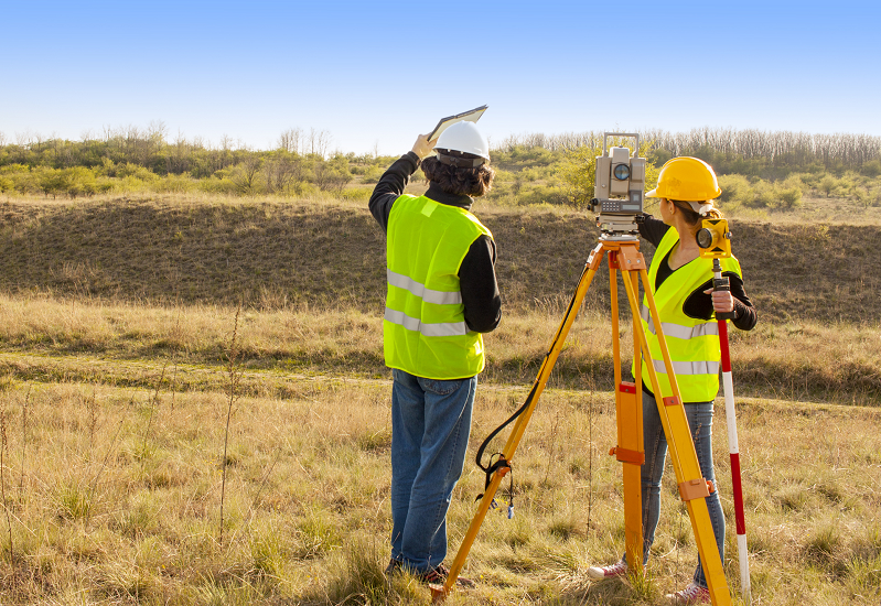 Two surveyors stand outside in a field with their equipment.