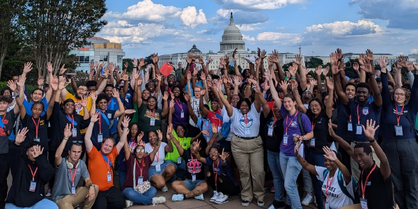 Dozens of young people gather for a rooftop group photo in front of the Capitol Building, raising their hands to the sky. 