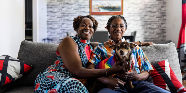 Two individuals smiling and sitting on a sofa in a cozy living room, holding a small dog wearing a rainbow bandana. The room is decorated with a brick-patterned wall and modern furnishings.