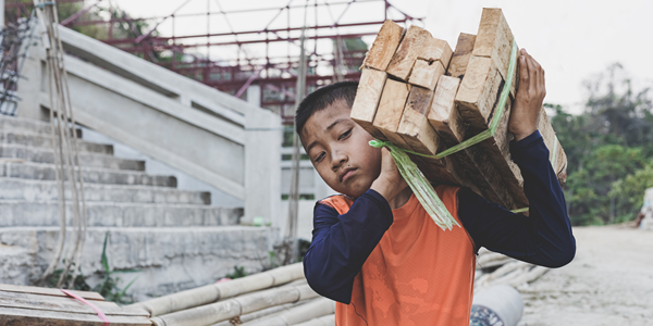 A boy carries a bundle of wood planks at a construction site.