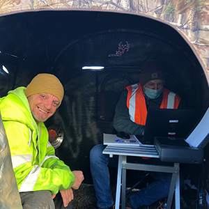 Two men in reflective jackets consult a computer in a makeshift shelter