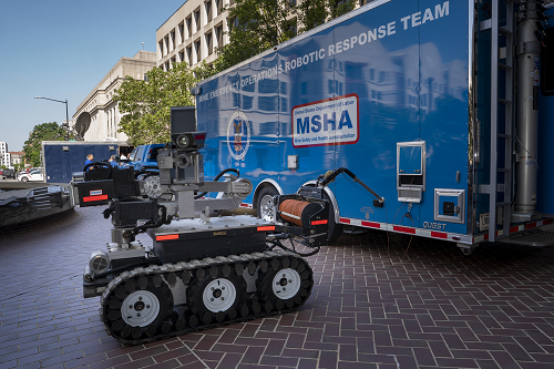 A robot with tracks like a bulldozer and a variety of mechanical parts sits outside a large blue trailer with MSHA's logo and "Mine Emergency Operations Robotic Response Team" 