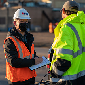 Two men in reflective safety jackets - one wearing a WHD hardhat and carrying a clipboard - chat outside