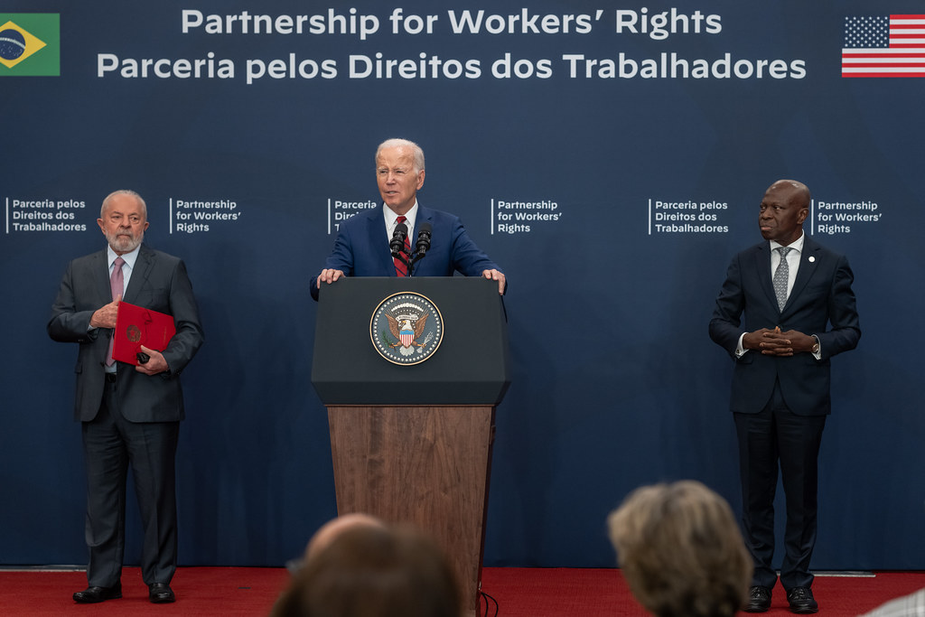 President Biden stands at a podium with Brazilian President Lula and ILO Director-General Houngbo by his side, the words "Partnership for Workers' Rights" written in English and in Portuguese above him.