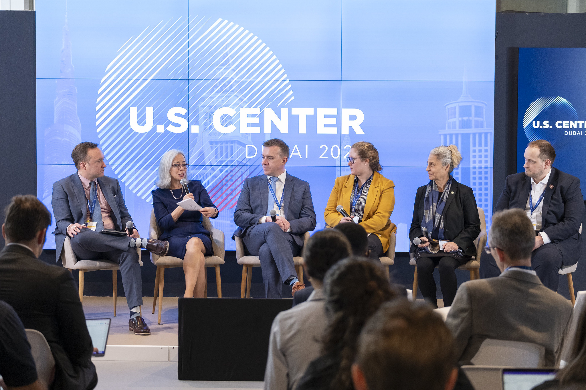Thea Lee sitting with a microphone on stage in a panel with five other people, the words "U.S. Center" illuminated on a panel behind her.
