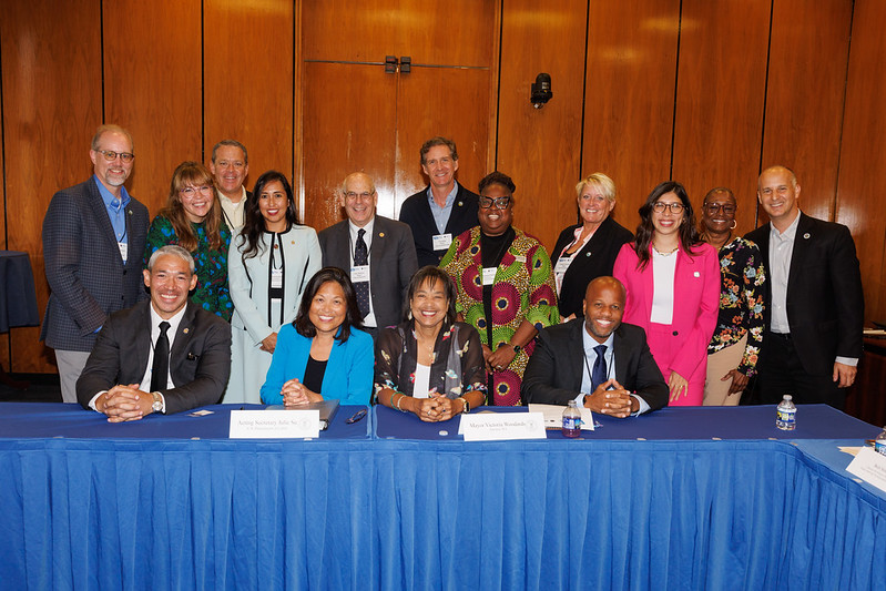 15 adults, all professionally dressed, gather for a group photo behind a conference table in a room with wood paneling. 