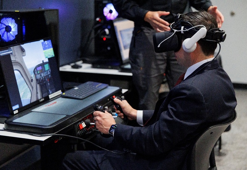 A man in a suit uses a virtual reality headset and aircraft controls as part of a training simulation.
