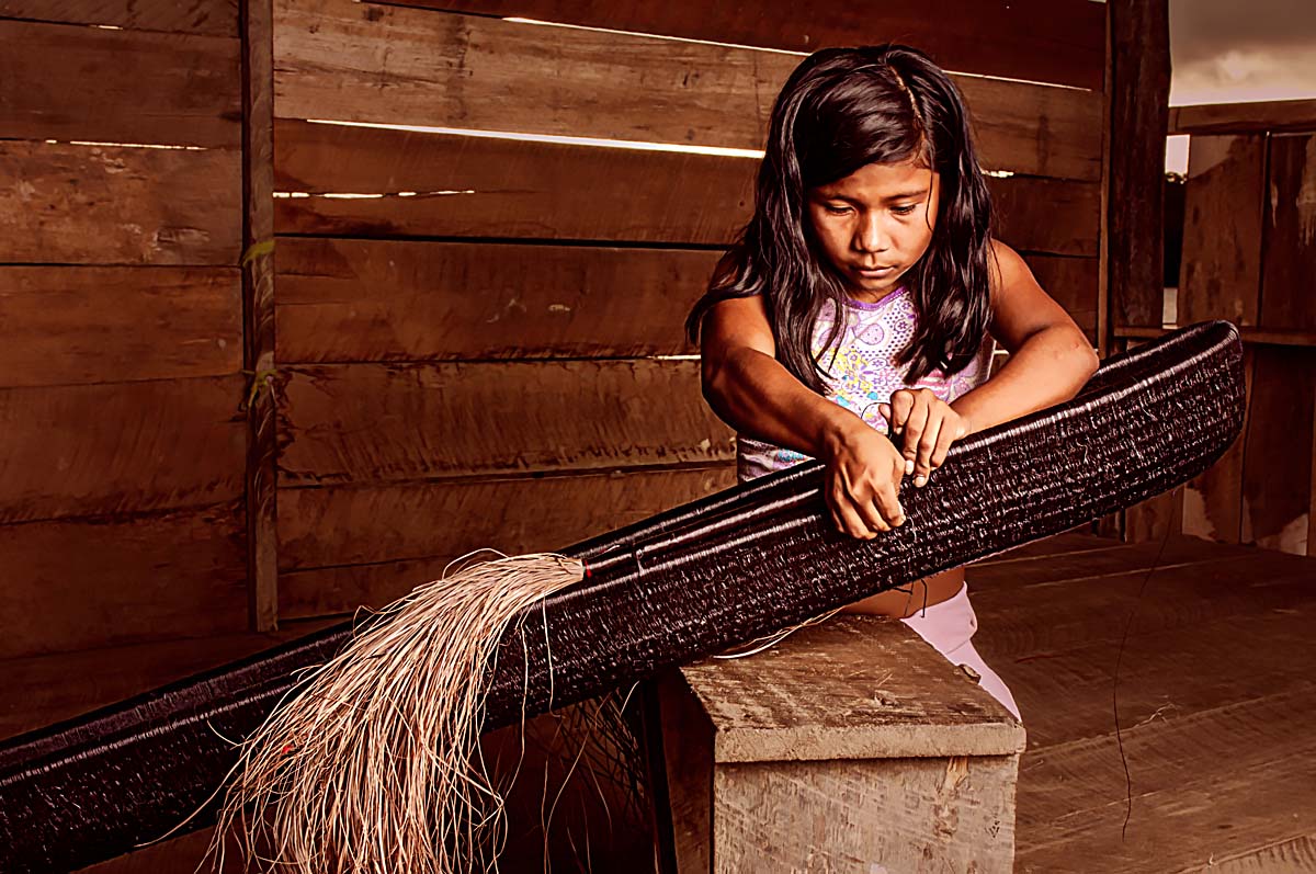A young girl weaves an oblong object on a work bench.