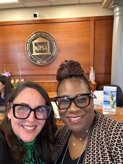 Dos mujeres con gafas negras sonríen para una selfie. El logotipo de la NJPSA es visible en la pared del fondo.