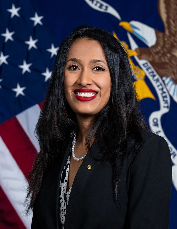 Image of a brown woman with long black hair who is smiling in front of the American and DOL flags.