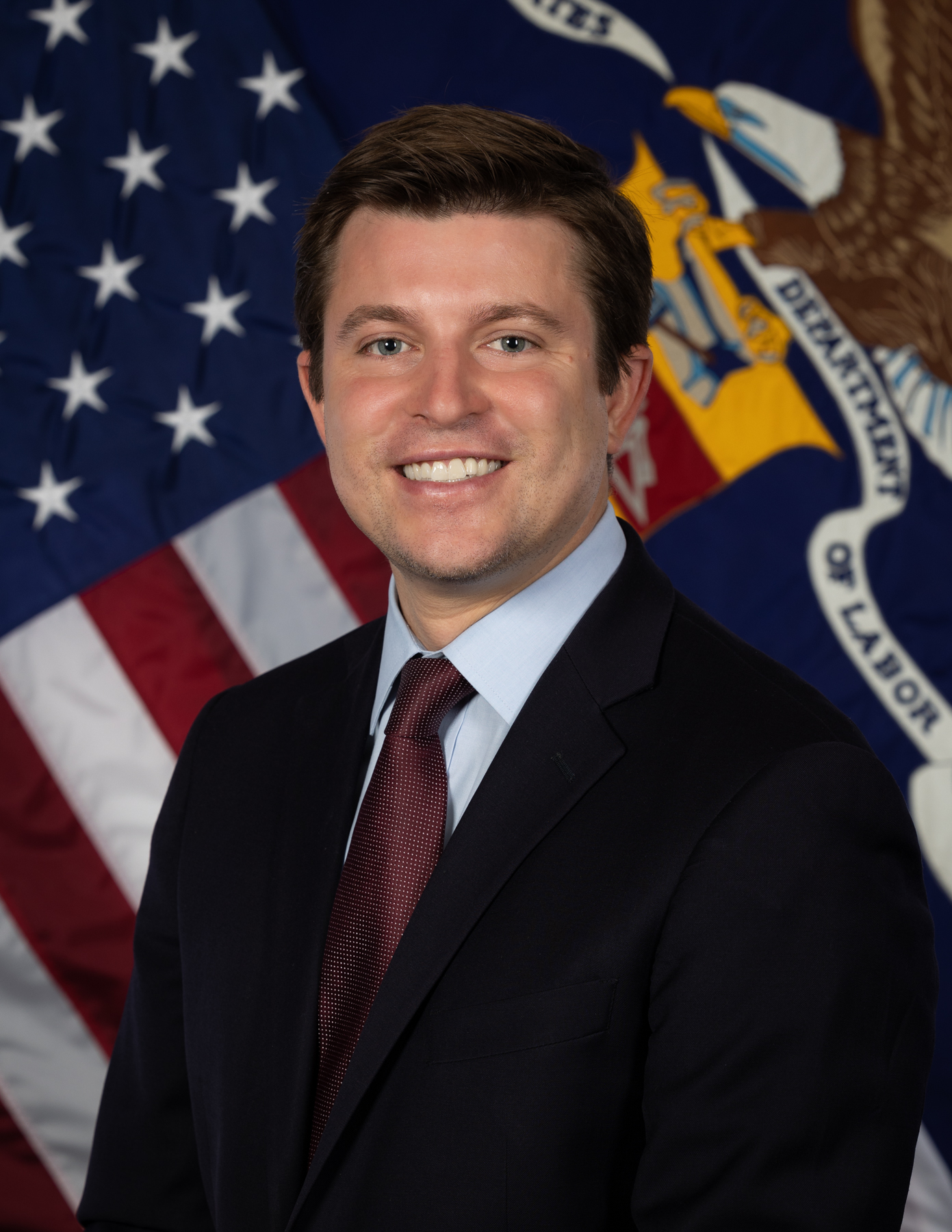 Official portrait of Brent Parton shows a man in business jacket smiling in front of the U.S. and Department of Labor flags.