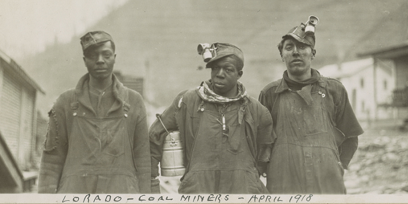 Three Black miners stand above ground, wearing coveralls and covered in coal dust.