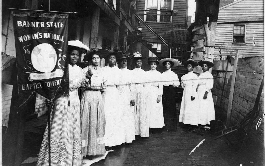 Nine African-American women posed, standing, full length, with suffragist Nannie Burroughs holding a banner reading, "Banner State Woman's National Baptist Convention