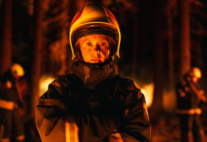 A female firefighter wearing her uniform and protective gear battling a fire in a smoky forest.