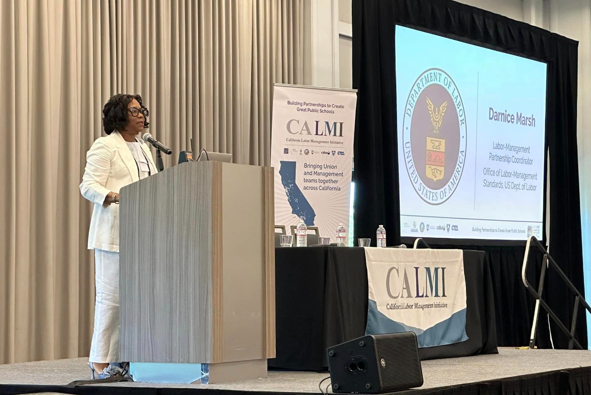 A woman wearing a white suit speaks at a podium. In the background there are signs for CALMI and a large screen showing her presentation.