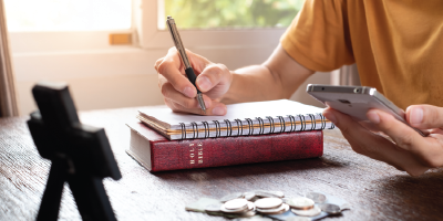 A person takes notes while using a calculator. A bible and cross are visible on the table.