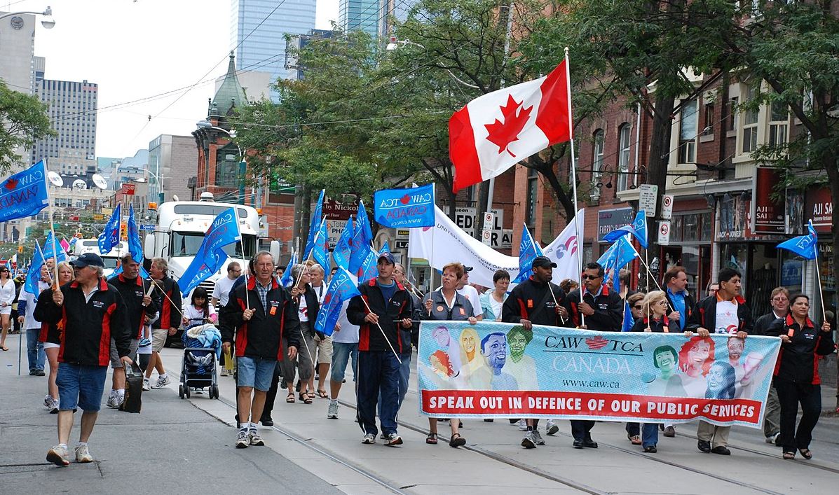 Picture of a large procession of workers marching down a street carrying Canadian flags and signs in support of workers