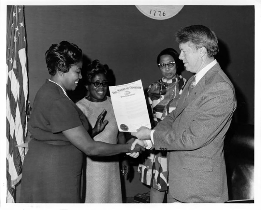 President Carter shakes Dorothy Bolden's hand and hands her a large piece of paper. Two other Black women look on in the background.