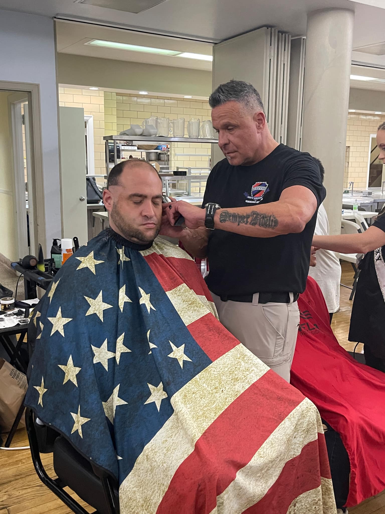 Chuck Ondo gives a man a haircut. The man in the barber chair has a beard and the smock covering him is a stars-and-stripes design.