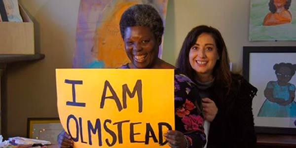 A smiling Black woman with short hair holds a sign reading 'I am Olmstead.' Paintings covers the walls behind her.