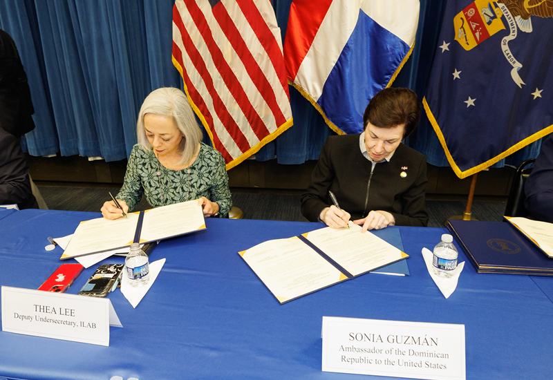 Dalawang babae ang nakaupo sa mesa na lumalagda ng mga dokumento. Nasa likod nila ang mga watawat ng Estados Unidos, Dominican Republic at Departamento ng Paggawa (Department of Labor) ng U.S.