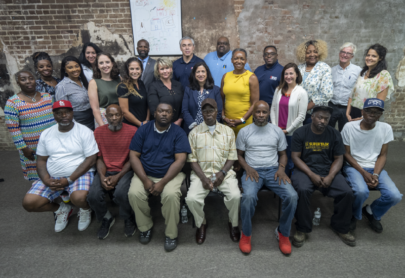 About two dozen people pose for a group photo in front of a brick wall.