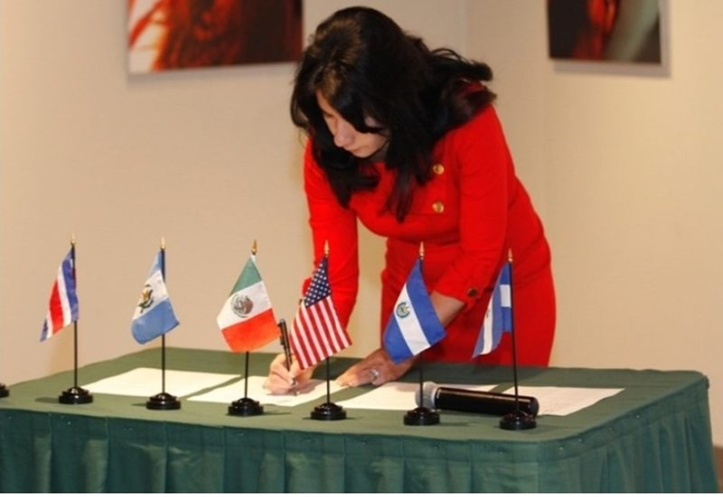 A woman in a bright red business suit signs a document on a table that has the flags of the United States, Mexico and several other countries.