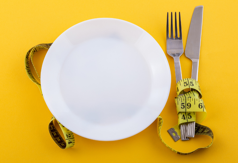 A white plate sitting next to silverware with yellow measuring tape wrapped around it on a yellow background.