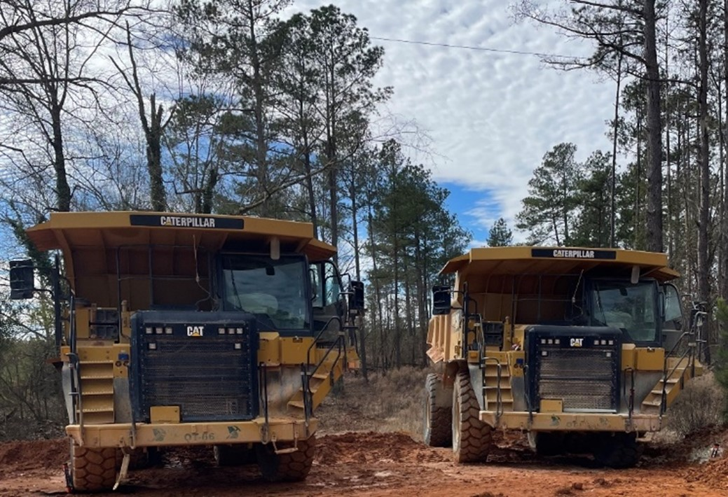 Two large dump trucks are parked in a wooded area with overhead power lines visible above them.