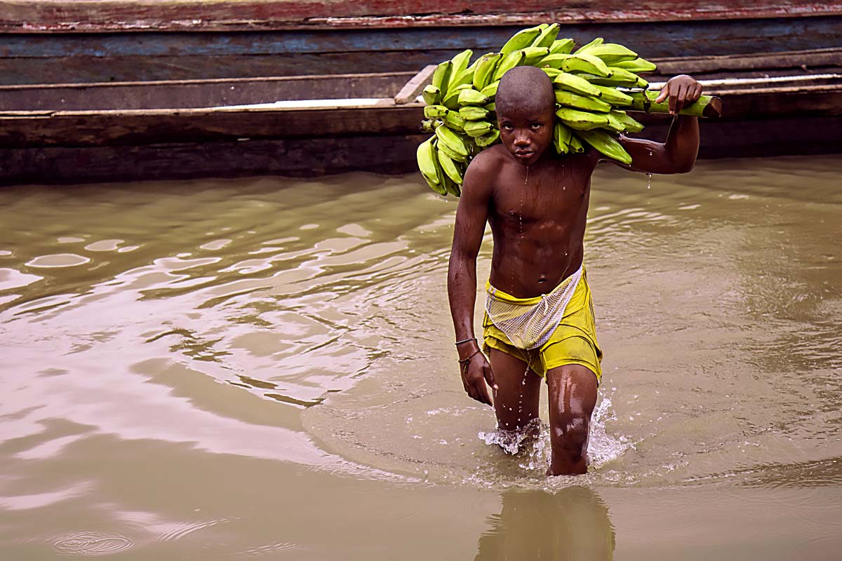 A young boy carries a stem of fruits on his back across the water.