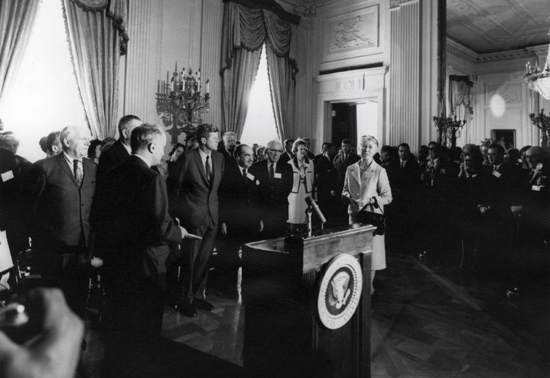 Black-and-white photo. Esther Peterson, President John F. Kennedy and dozens of others others stand in the White House. A podium is in the foreground.