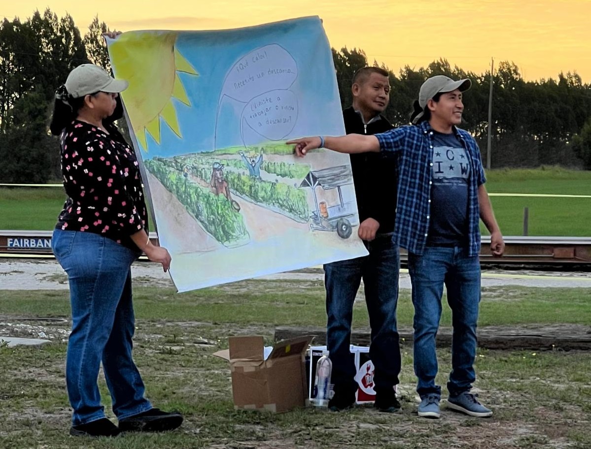 Standing outside at sunset, a worker points to a painted picture of farmland being held up by two colleagues.