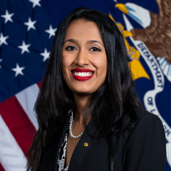 Professional head shot of a woman with dark hair smiling in front of a Labor Department flag.