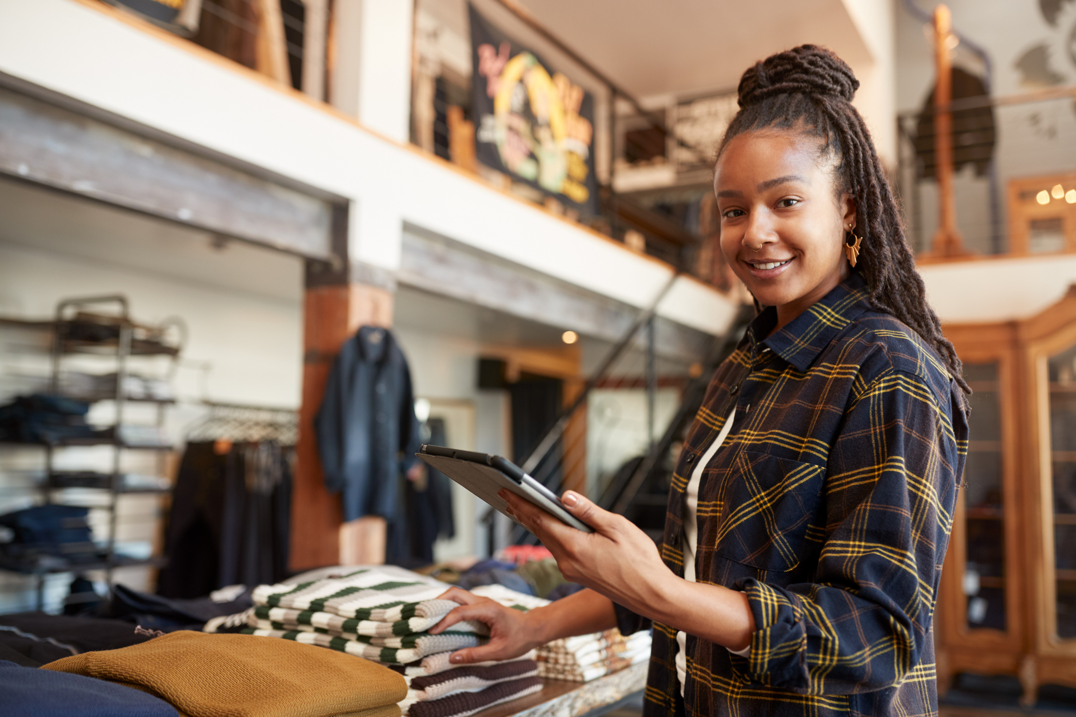 A woman holding a tablet folds sweaters on a table in a shop.