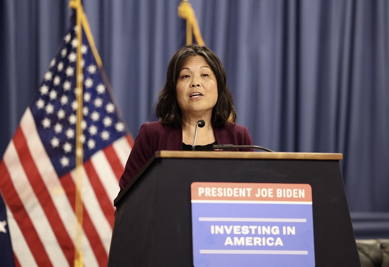 Acting Secretary Julie Su speaks at the Department of Labor's "Good Jobs, Great Cities Academy" launch event. She is standing behind a podium with a sign that says "President Joe Biden: Investing in America."