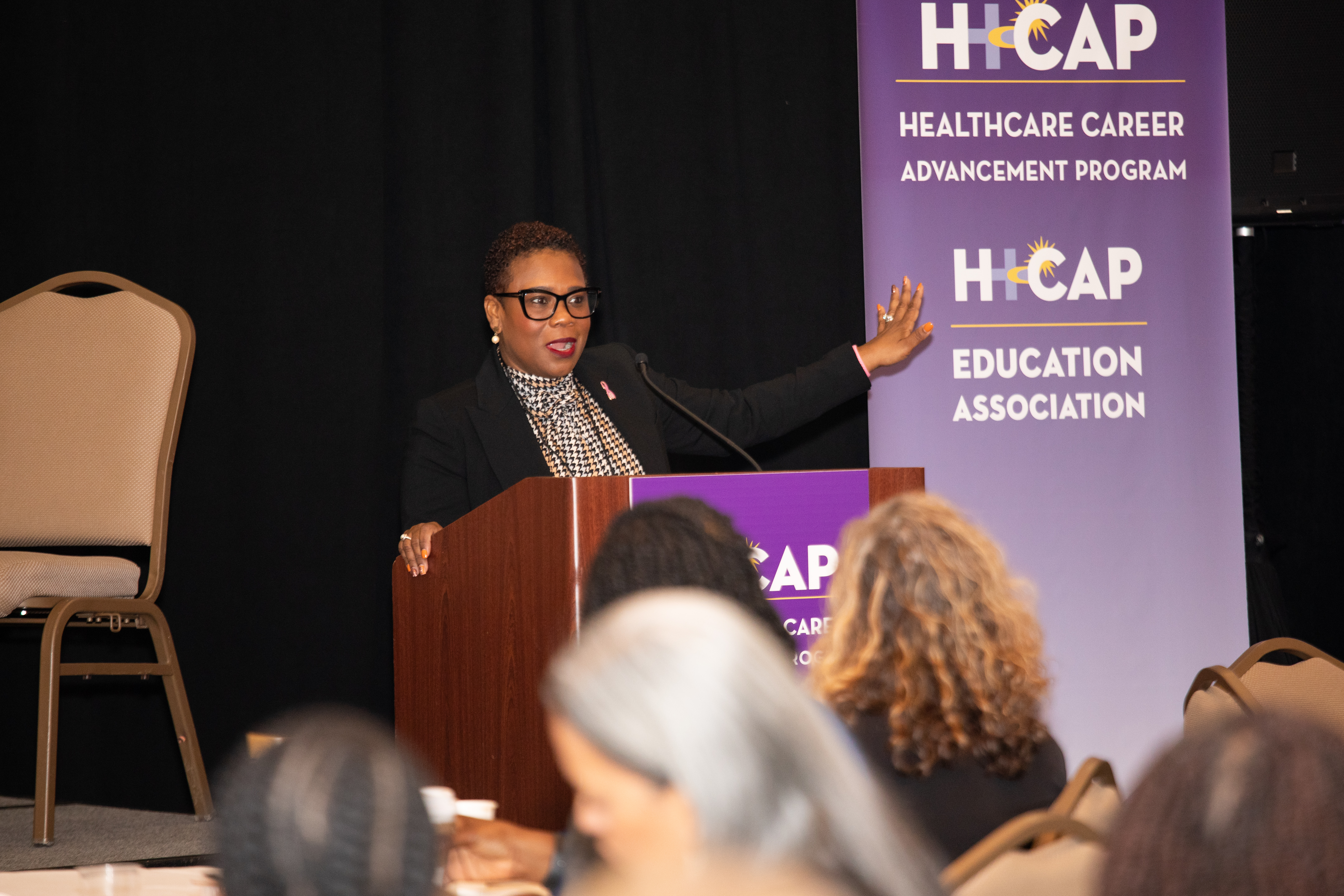 A woman in a dark jacket and glasses speaks at a podium. A banner in the background reads "H-CAP Healthcare Career Advancement Program. H-CAP Education Association."