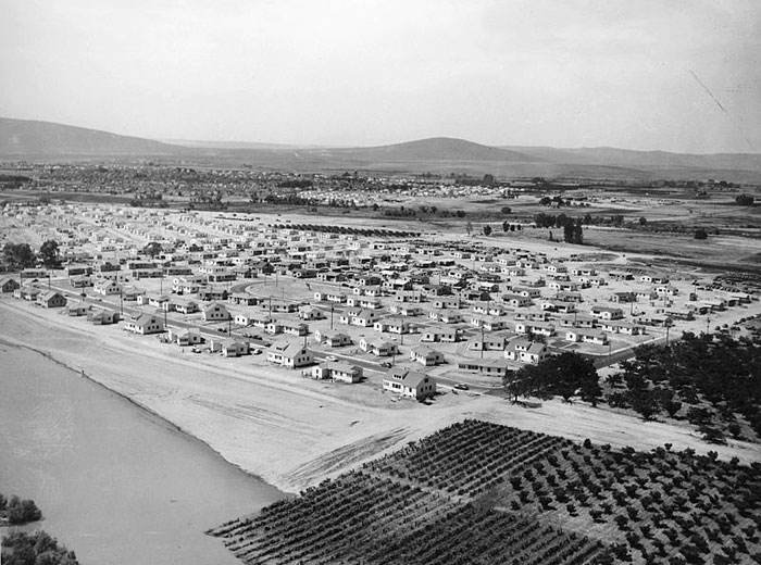 Black and white aerial photograph of a cluster of mostly small houses on a plain, with gently sloping hills in the distance.