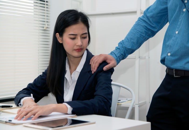 Image description: A woman sitting at a desk looks apprehensively at the hand of a standing man that’s placed on her shoulder. Both are wearing office attire.
