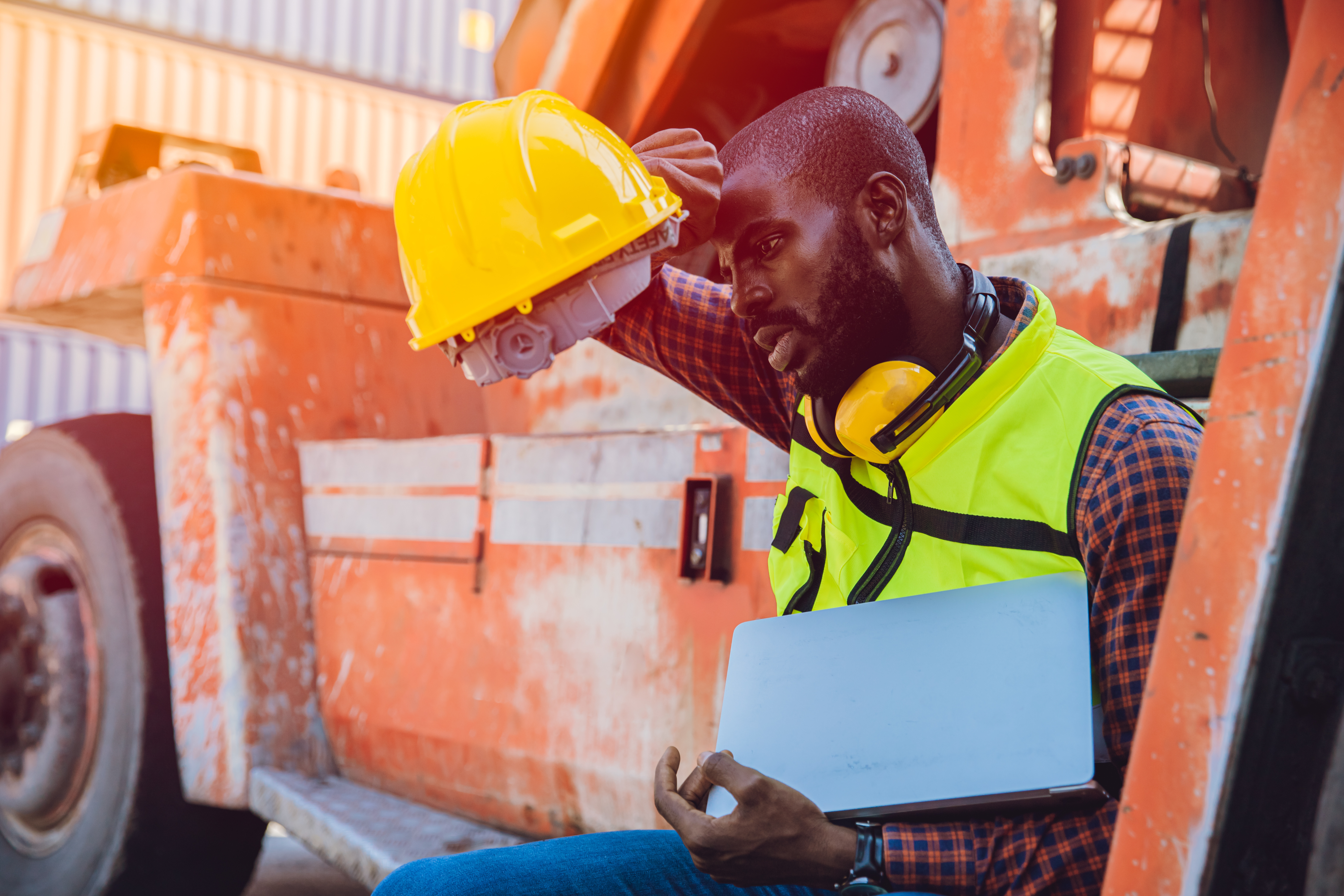 A worker sits down on a work vehicle to cool down. They are wiping sweat off of their forehead.