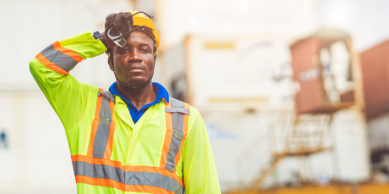 A worker wearing reflective clothing stands outside in the heat, wiping his brow