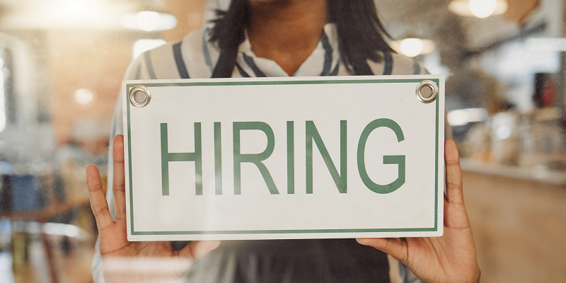 A female worker in a coffee shop wearing an apron hangs a "hiring" sign on the window.