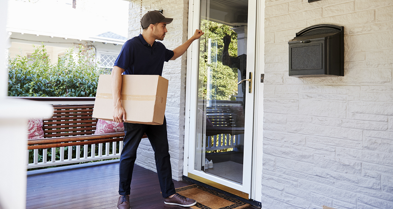 A delivery worker carries a large cardboard box to the front porch of a home, and knocks on the door.