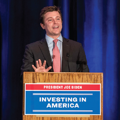 A man in a suit speaks at a podium labeled "President Joe Biden - Investing in America"