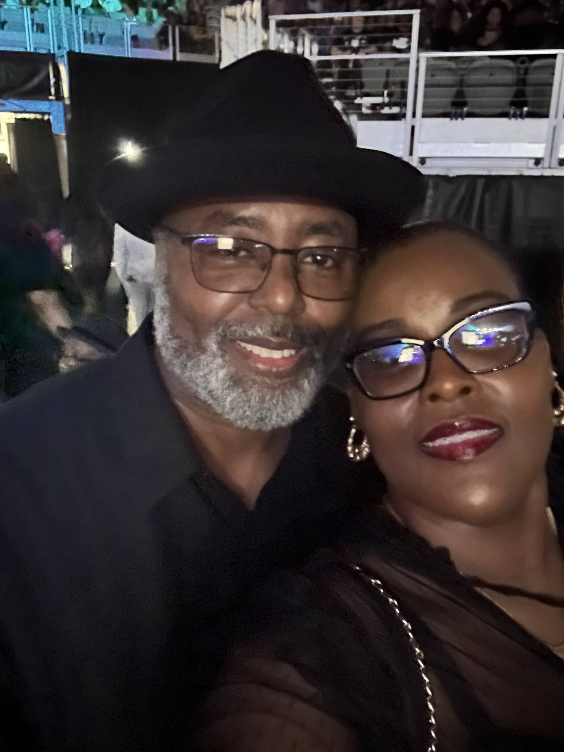 James and Pamela Jackson smile at the camera. Both are wearing glasses, and James is wearing a hat. They are at a concert venue. 