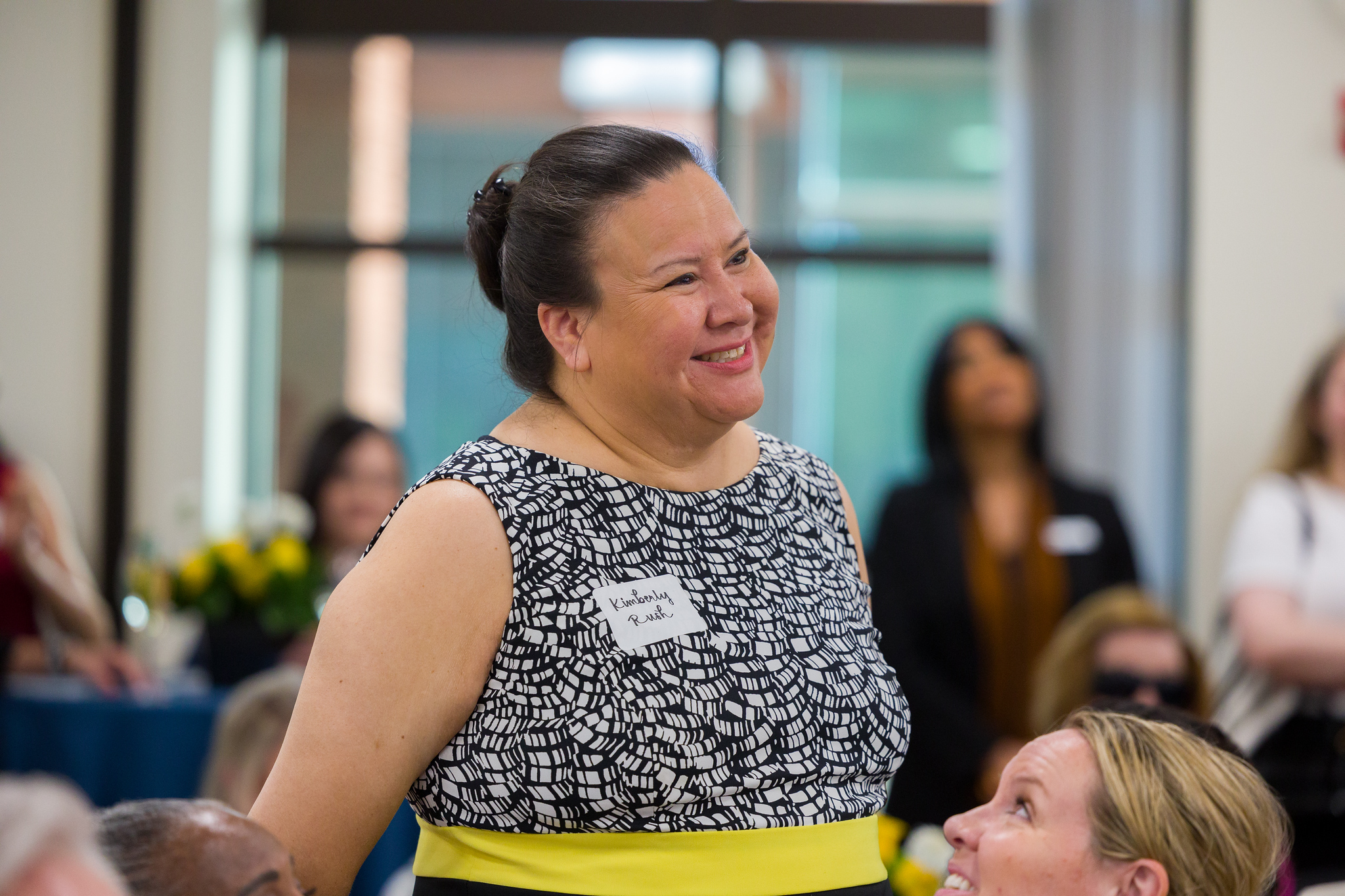 A woman wearing a nametag that says “Kimberly Rush” stands in a conference room, surrounded by other women. She is smiling. 